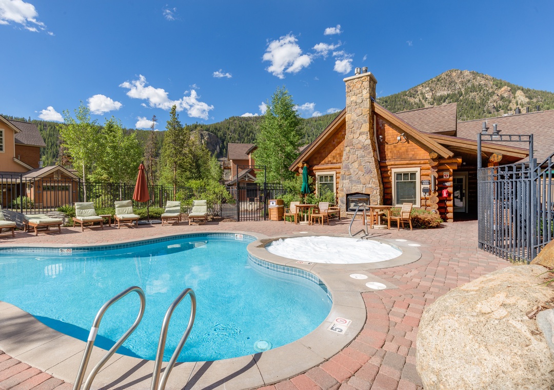 Resort-style pool area surrounded by mountain scenery, featuring a stone fireplace clubhouse and relaxing outdoor seating with dramatic peaks as backdrop.