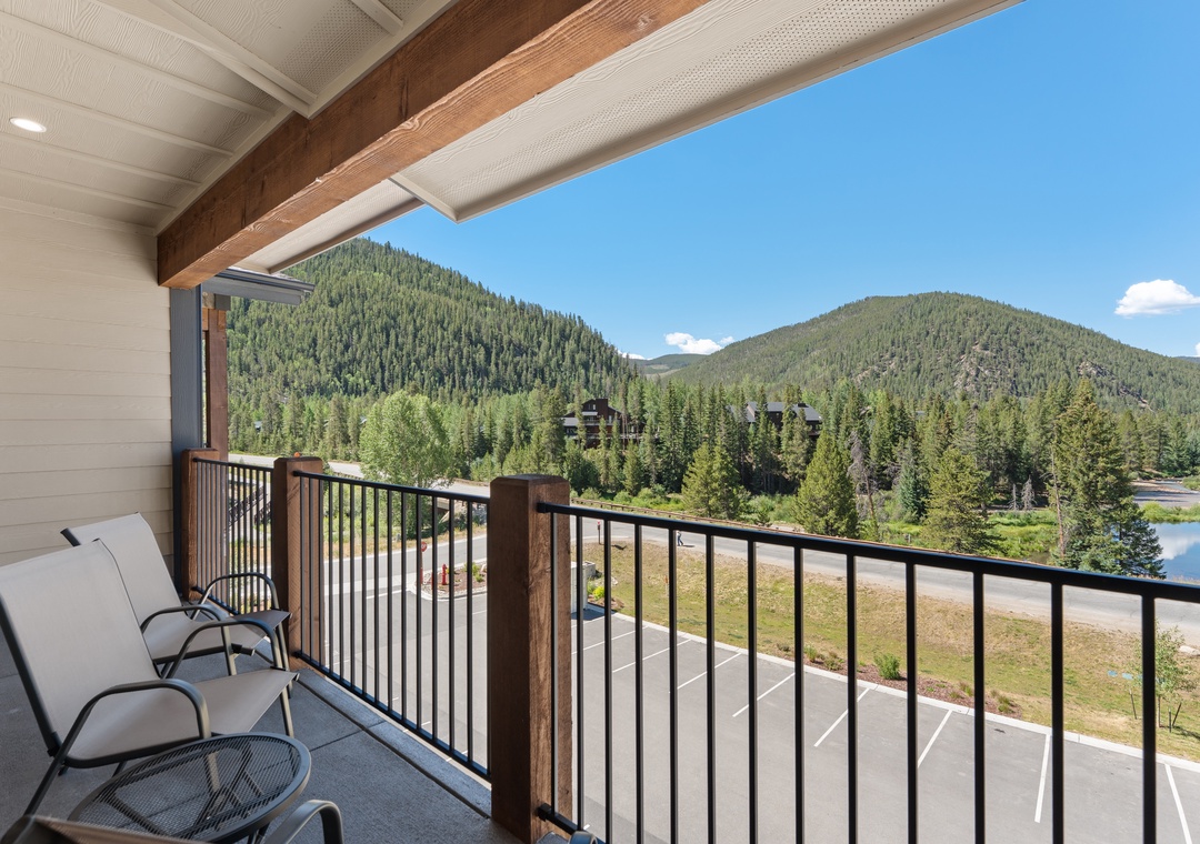 Your private balcony overlooks a serene natural landscape where lush pine forests meet tranquil waters and rolling mountain peaks under clear blue skies.