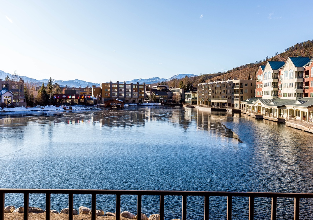 Mountain town waterfront with colorful buildings reflecting in calm waters, surrounded by scenic peaks and forested hills.