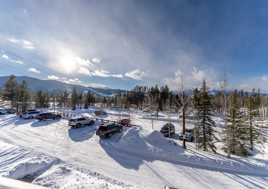 Expansive mountain vista with snow-covered parking area surrounded by evergreen forest and distant peaks under dramatic winter sky.