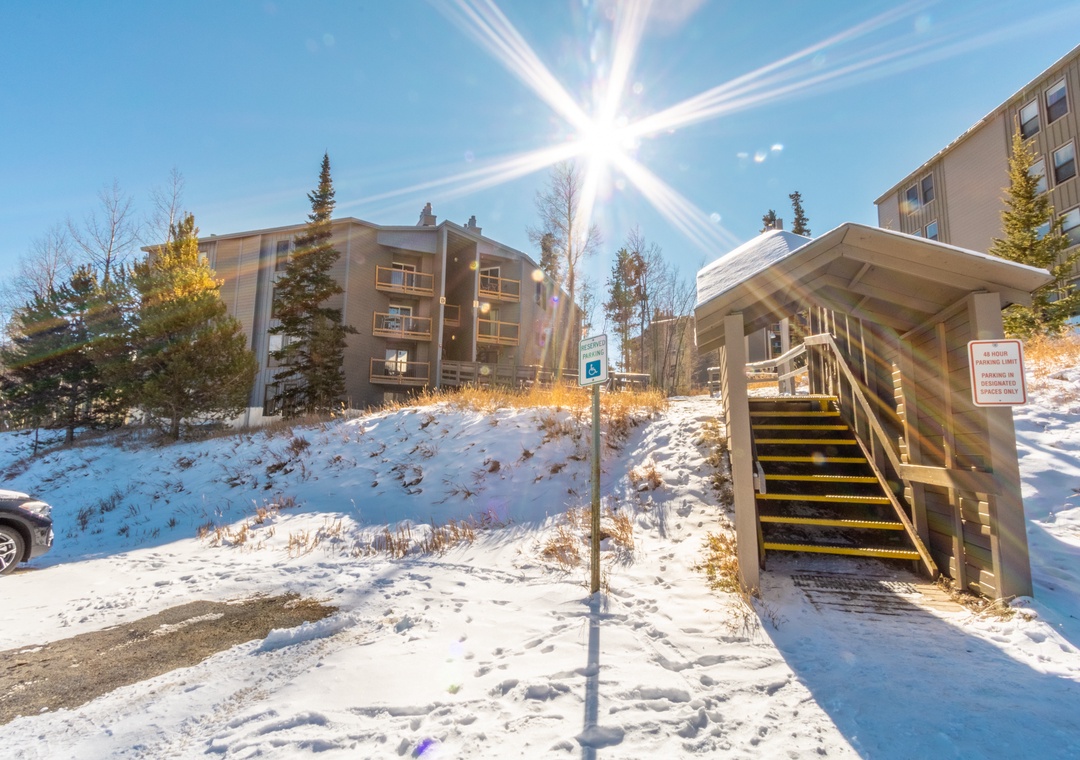 Snow-covered property complex with modern apartment buildings surrounded by evergreen trees under bright winter sunlight.