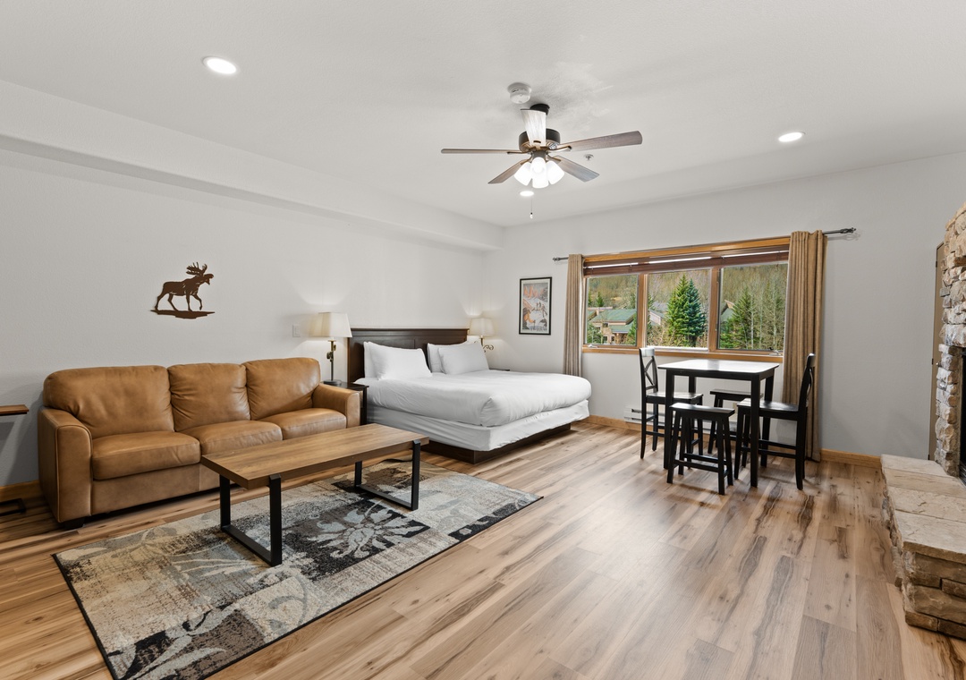 Your spacious studio welcomes you with warm wood floors, a cozy tan sofa, and a plush bed near bright windows overlooking mountain trees and greenery.
