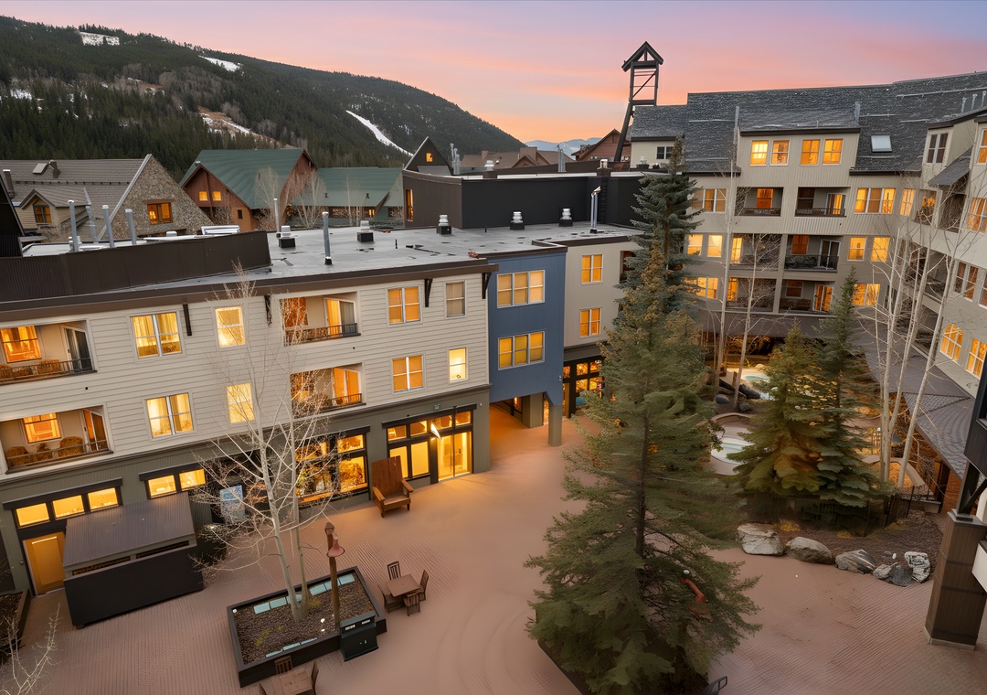 A mountain village courtyard framed by modern multi-level buildings, with ski slopes visible on the peaks and warm windows glowing at dusk.
