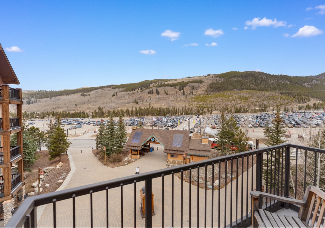 A ski resort base lodge sits below rolling mountains with extensive parking areas and forest surroundings under clear blue skies.