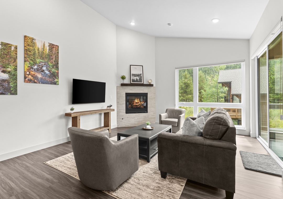 Settle into your cozy living room where modern comfort meets natural warmth. The glowing fireplace creates an inviting ambiance while you unwind on plush seating and enjoy serene views of the surrounding greenery through expansive windows.