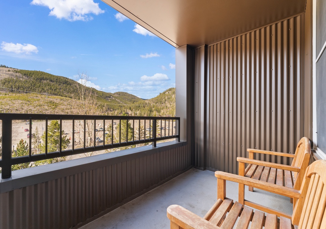 The covered balcony features two wooden chairs facing a stunning mountain vista framed by forested hills and open sky—a peaceful retreat on the property's exterior.