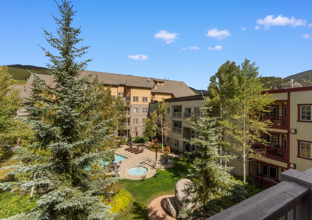 Mountain resort complex with pool and hot tub nestled among evergreen trees under clear blue skies.