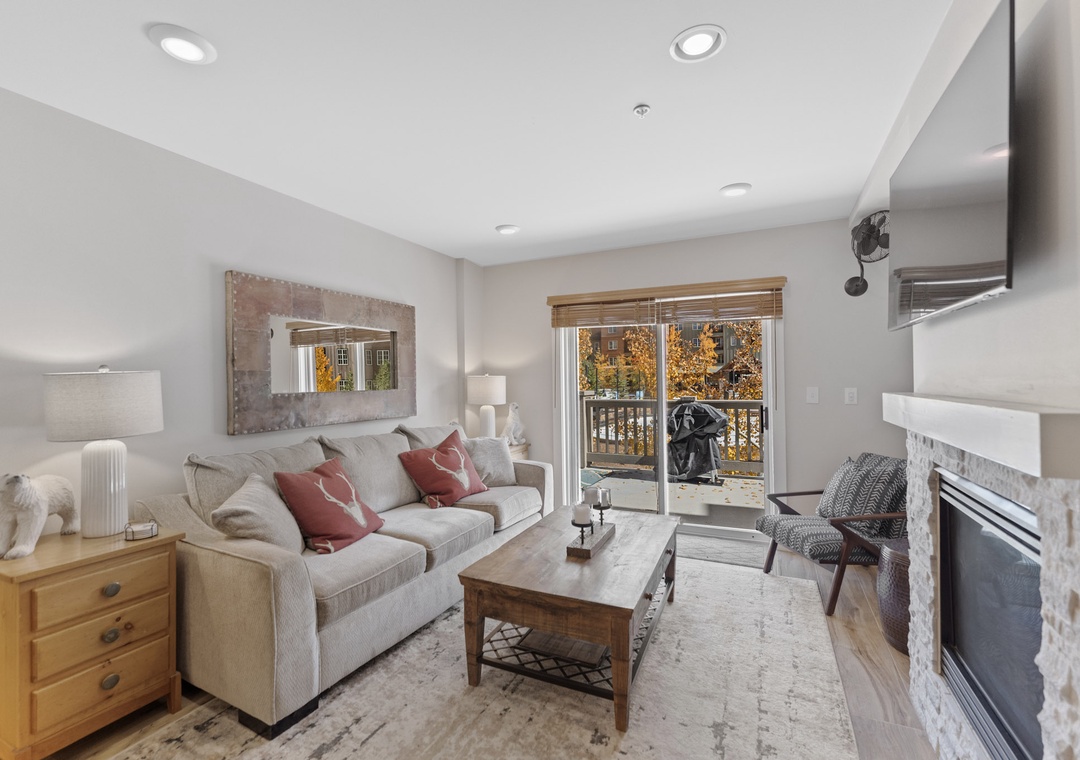 Relax in your cozy living room with plush seating, warm fireplace, and peaceful balcony views of autumn foliage.