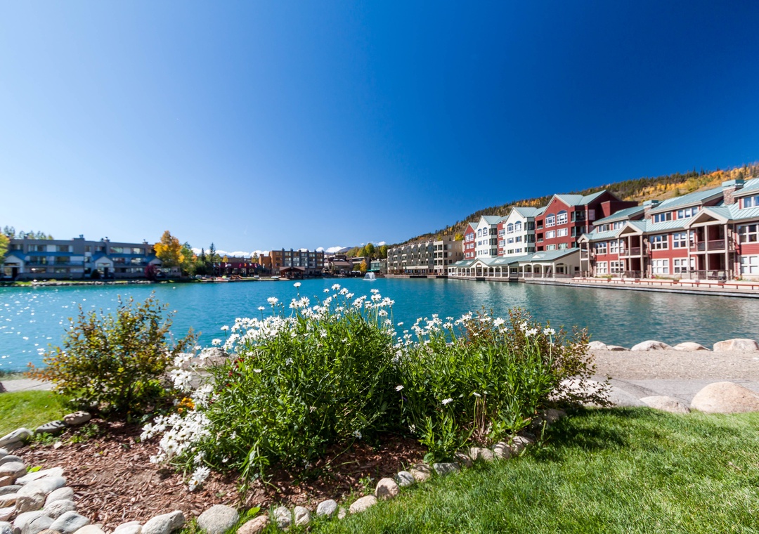 Charming lakeside village with colorful buildings reflecting in pristine blue waters, surrounded by autumn foliage and manicured gardens.