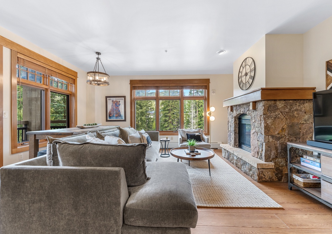 Sink into plush gray sofas by your cozy stone fireplace, where crackling warmth meets forest views through expansive windows.