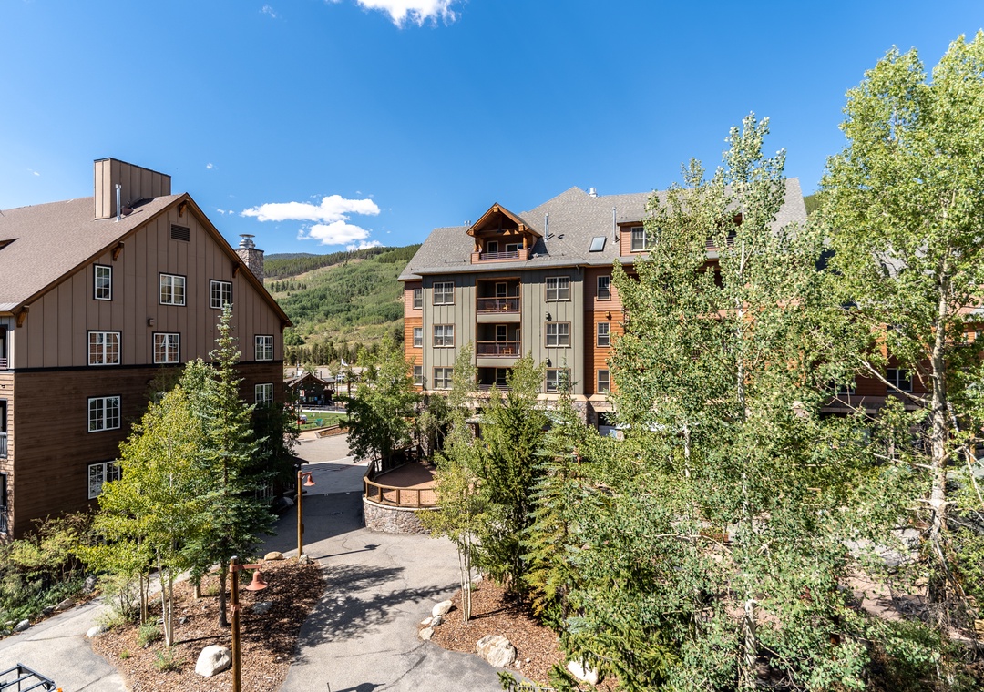 Mountain resort buildings nestled among mature trees with forested hills rising in the background under a bright blue sky.