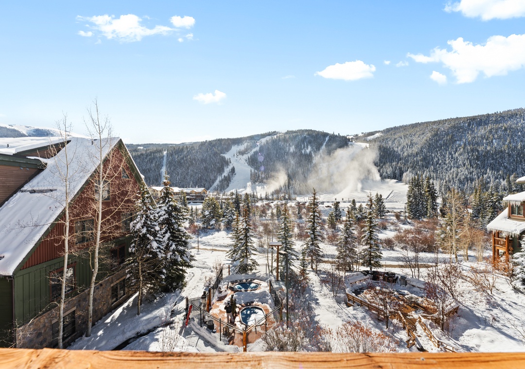 Stunning mountain ski resort view with snow-covered slopes and winter village below, framed by evergreen forests under bright blue skies.