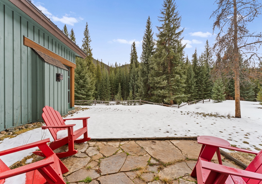 Charming mountain cabin with red Adirondack chairs on stone patio, surrounded by pristine forest and snow-dusted grounds.
