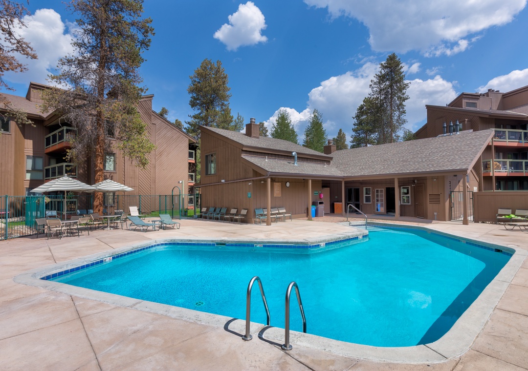 Resort-style pool area with sparkling blue water, surrounded by comfortable loungers and shaded seating under mature pines.