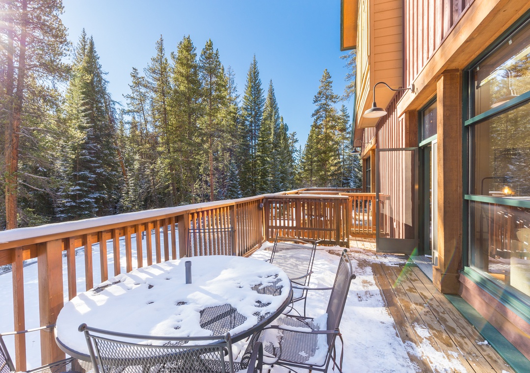 Snow-dusted balcony with outdoor dining table surrounded by towering evergreens and winter mountain scenery.