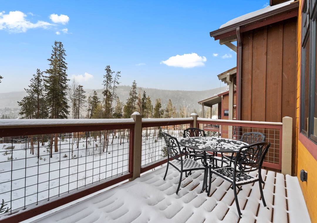 Snow-covered balcony with mountain views and outdoor dining furniture surrounded by winter forest landscape.