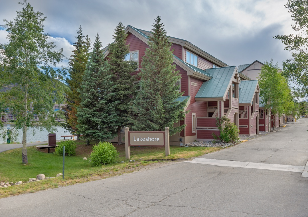Lakeshore property entrance showcases distinctive red townhomes with green metal roofs nestled among mature evergreen and deciduous trees.