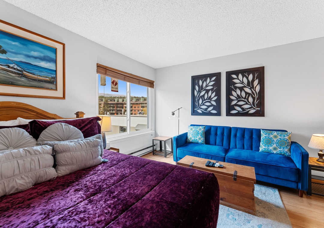 Relax in your bright bedroom featuring a plush queen bed, vibrant blue sofa, and charming city views from your window.