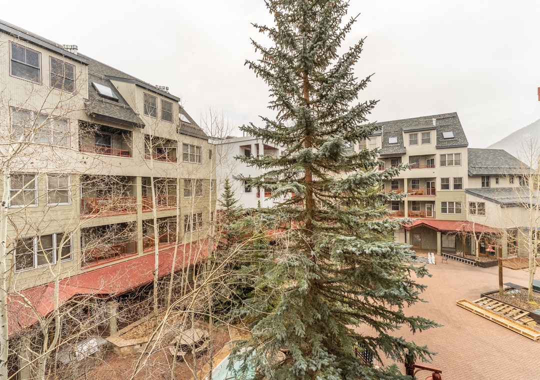 Multi-story residential buildings surround a central courtyard with mature evergreen trees and mountain backdrop.