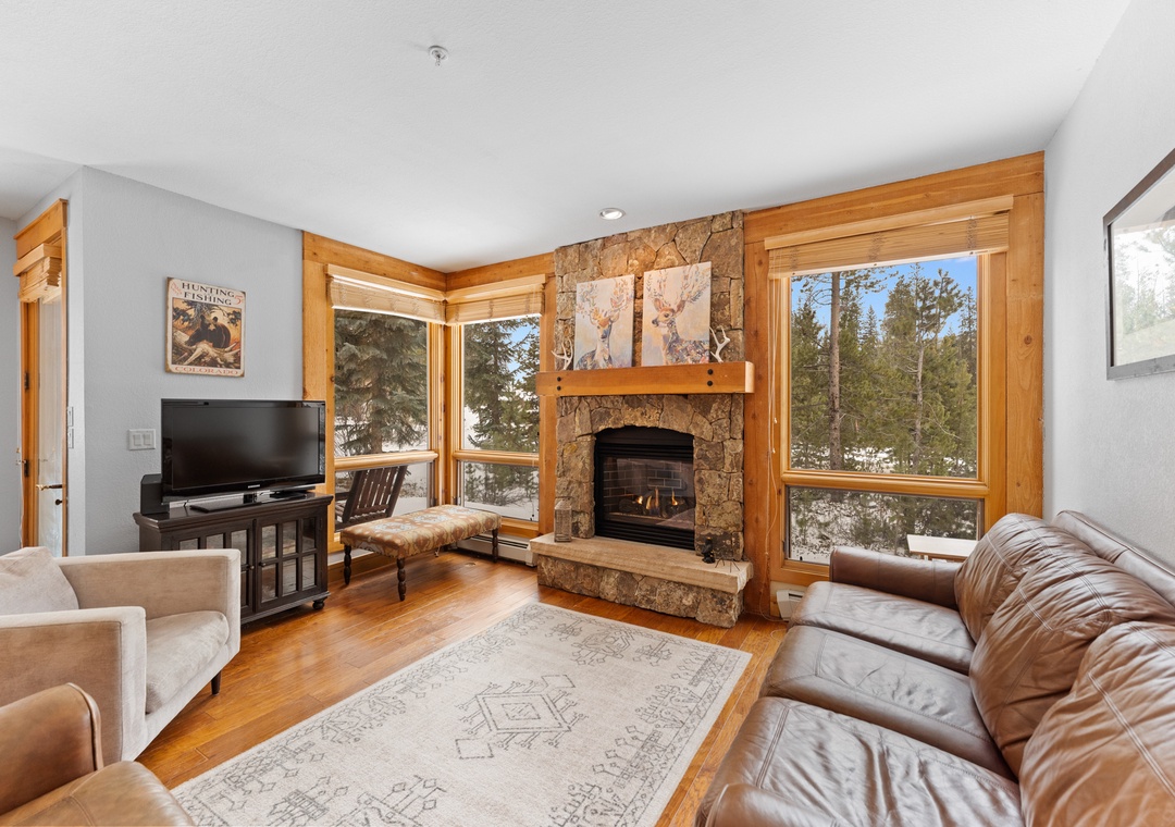 Cozy up in your mountain living room where a crackling stone fireplace meets panoramic forest views through expansive windows.