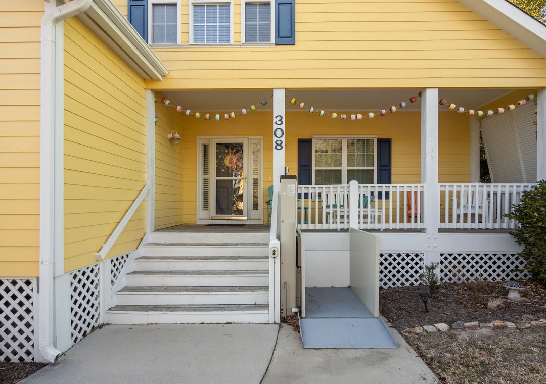 Charming yellow cottage with cheerful porch featuring festive string lights and accessible ramp entrance.