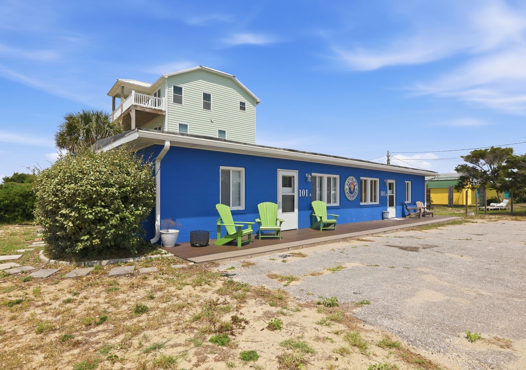 The vibrant blue building and neighboring green house create a colorful coastal arrival point, with numbered doors opening to your beachside escape and cheerful Adirondack chairs promising relaxation.