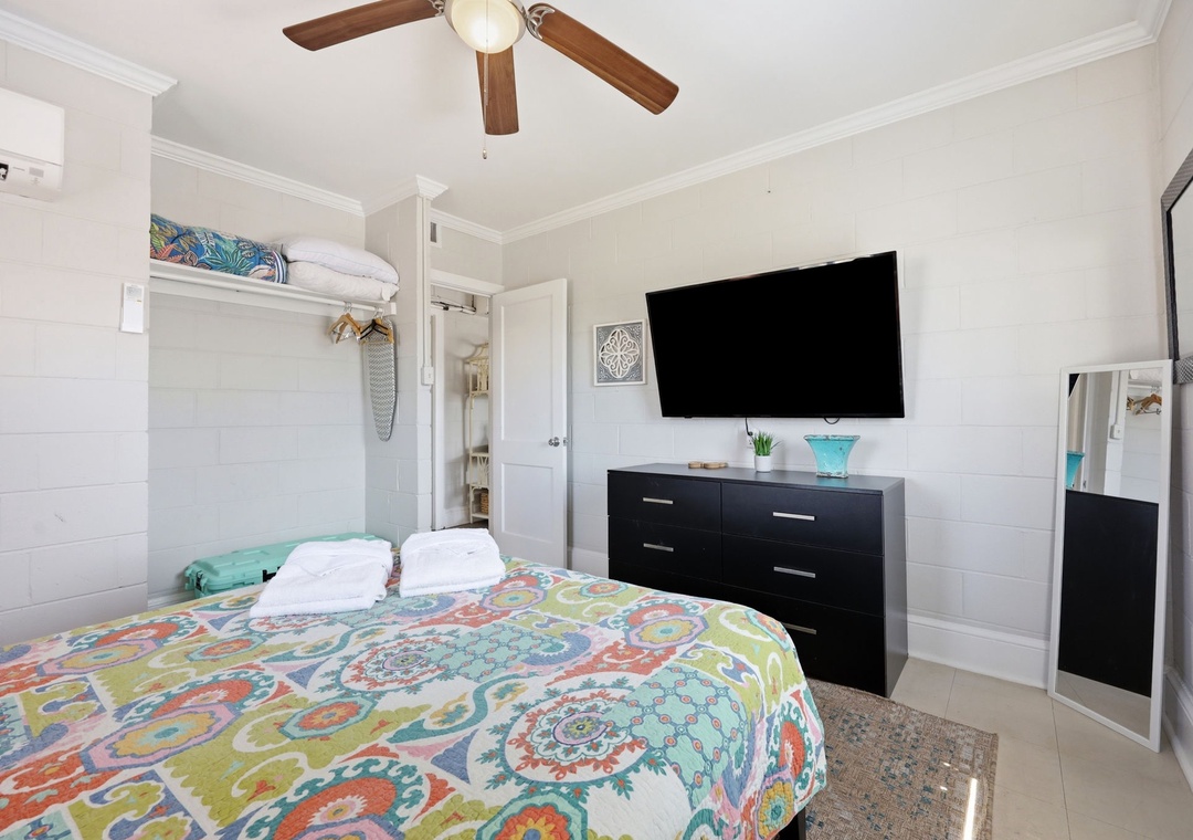 Relax in your bright, airy bedroom featuring a cozy bed with colorful linens, a wall-mounted TV for your entertainment, and a ceiling fan with climate control to keep you comfortable year-round.