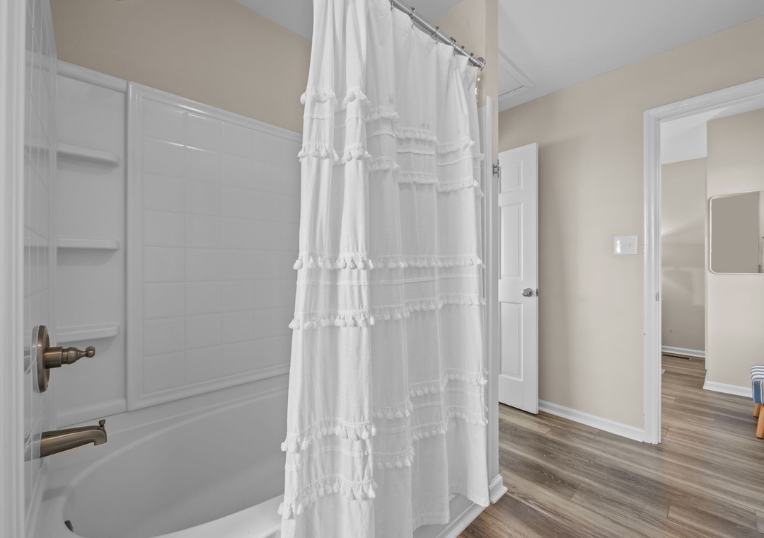 Step into your bright, clean bathroom featuring a spacious tub-shower combo with charming ruffled curtain and beautiful flooring throughout.