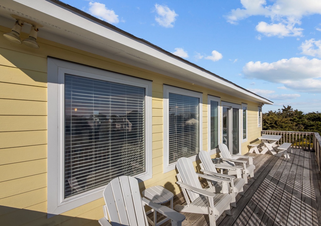 Cheerful yellow coastal cottage exterior featuring a spacious deck with classic Adirondack chairs overlooking surrounding greenery under bright blue skies.