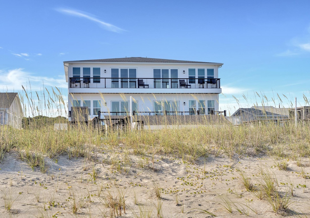 Beachfront property featuring direct sandy beach access with dunes and beach grasses leading to a multi-level coastal building with private balconies under clear blue skies.