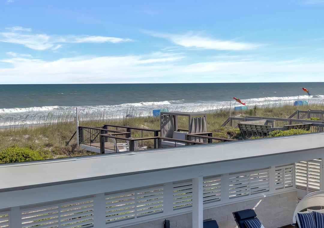 A sunny beachfront location with direct access to sandy shores. The property features a raised deck with ocean views, separated from the beach by dunes and a wooden walkway. The area offers a peaceful coastal retreat with convenient beach proximity.