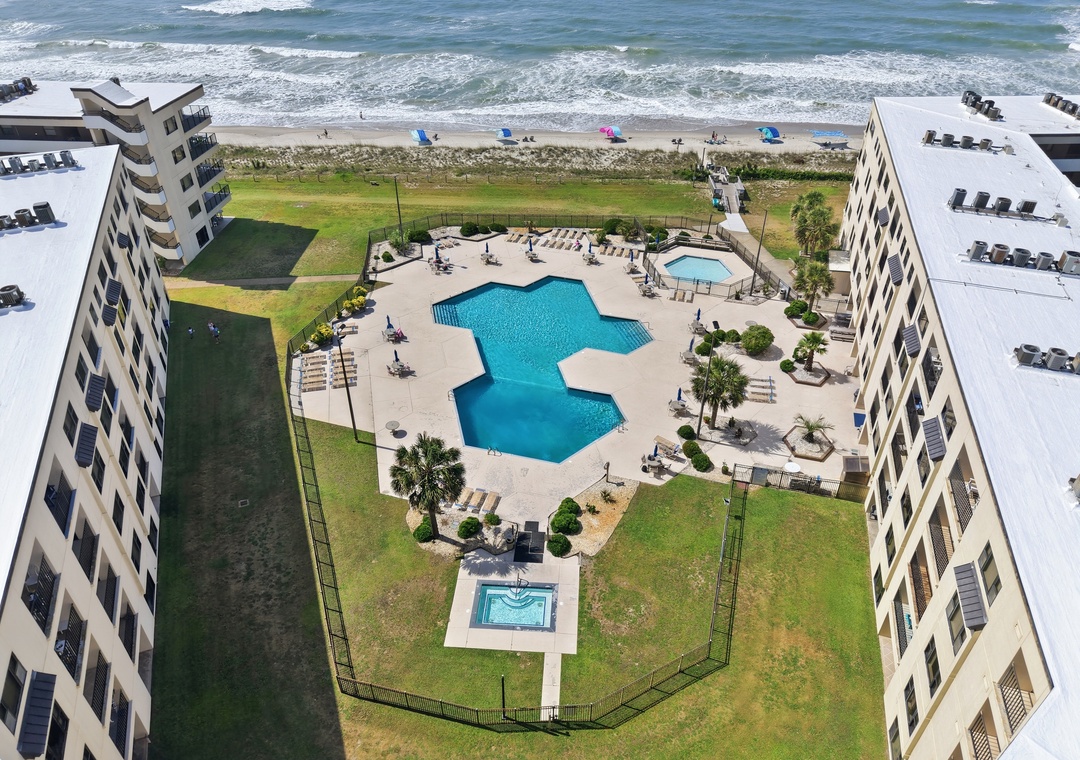 Aerial view of the beachfront property showcasing dual swimming pools, a hot tub, and lounge area nestled between residential buildings with direct beach access and ocean beyond.