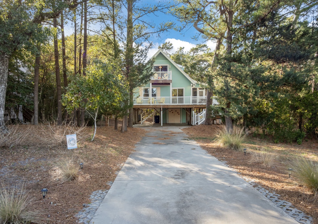 A charming coastal cottage nestled among tall pines, featuring multiple decks and a welcoming driveway leading to your peaceful forest retreat.