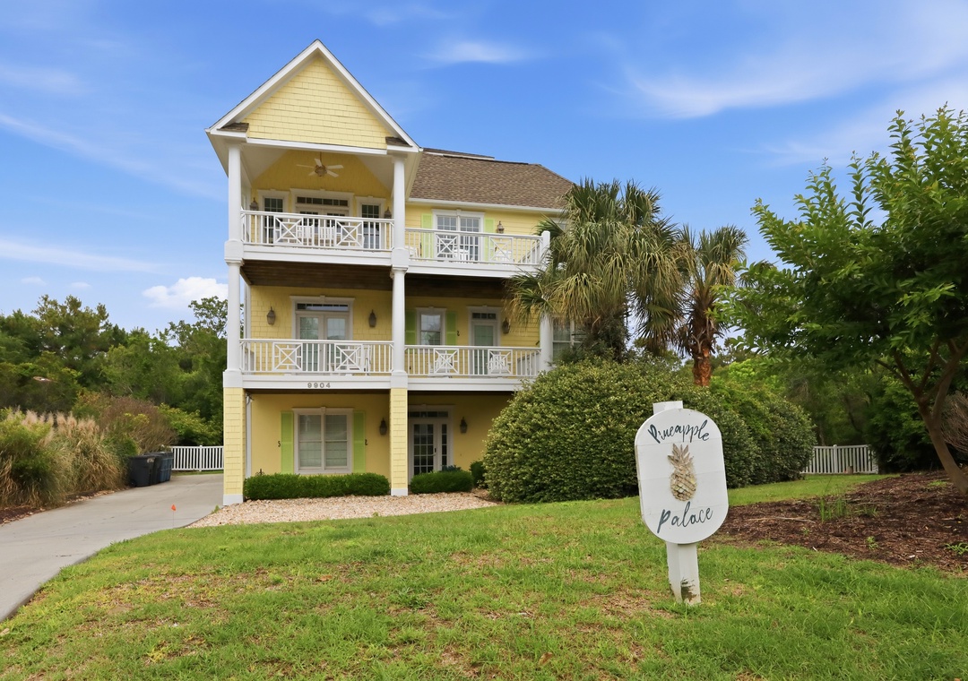 Pineapple Palace welcomes guests with cheerful yellow siding, multiple balconies, and lush coastal landscaping under clear blue skies—a bright, inviting arrival experience.