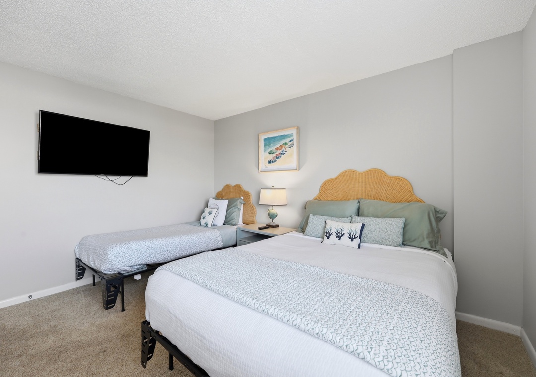 Your coastal-inspired retreat features two plush beds with woven rattan headboards, serene ocean-toned linens, and a wall-mounted TV for your entertainment. Drift off to the gentle seaside palette surrounding you.