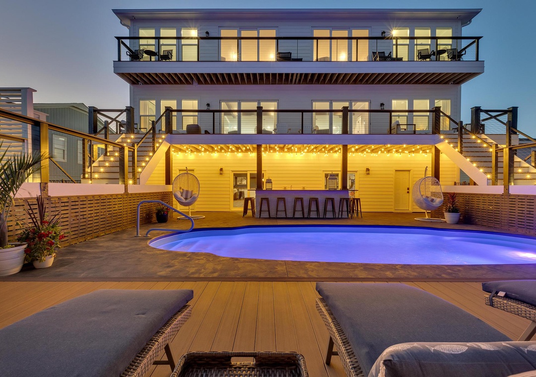 The modern three-story property features expansive balconies on each level, an illuminated pool with integrated bar seating, and an inviting outdoor entertainment area perfect for sunset gatherings.