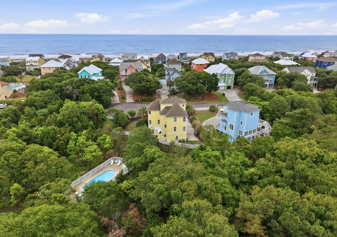Aerial view of a coastal vacation rental community featuring colorful beach homes nestled among lush green trees, with ocean views and a private pool on-site.