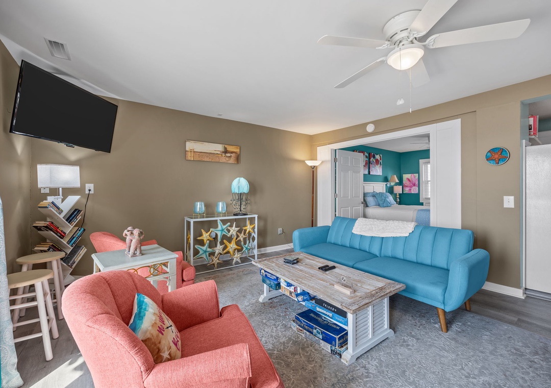 Unwind in your bright living room with plush coral and teal seating, ceiling fan comfort, and flat-screen entertainment after beach days.