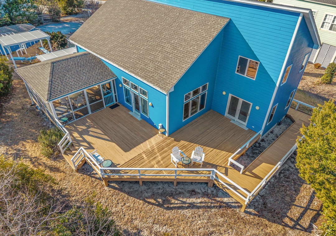 Modern blue beach house featuring expansive wraparound deck with Adirondack chairs, perfect for morning coffee and sunset views in peaceful residential setting.