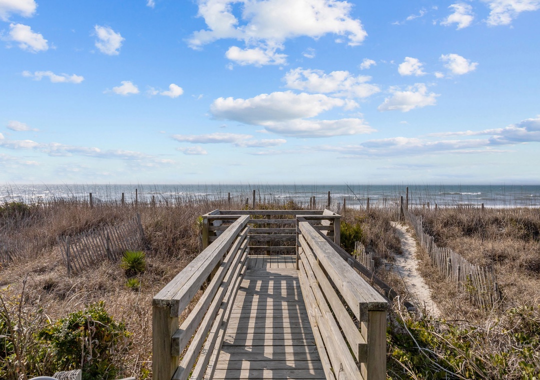 A wooden boardwalk leads through coastal dunes to the beach, with ocean views and blue skies creating a perfect pathway to relaxation.