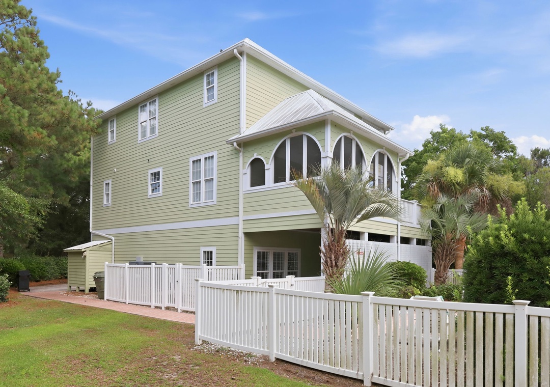 A charming three-story coastal home with soft green siding and white trim, featuring covered parking and a white picket fence, surrounded by lush palms and mature trees in a peaceful neighborhood setting.