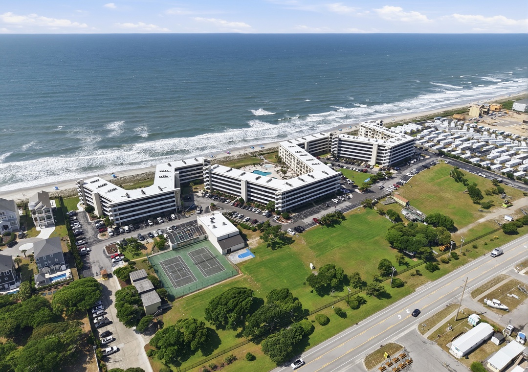 Aerial view of a beachfront resort complex featuring tennis courts and green spaces, nestled along a pristine sandy coastline with rolling waves and open ocean beyond.