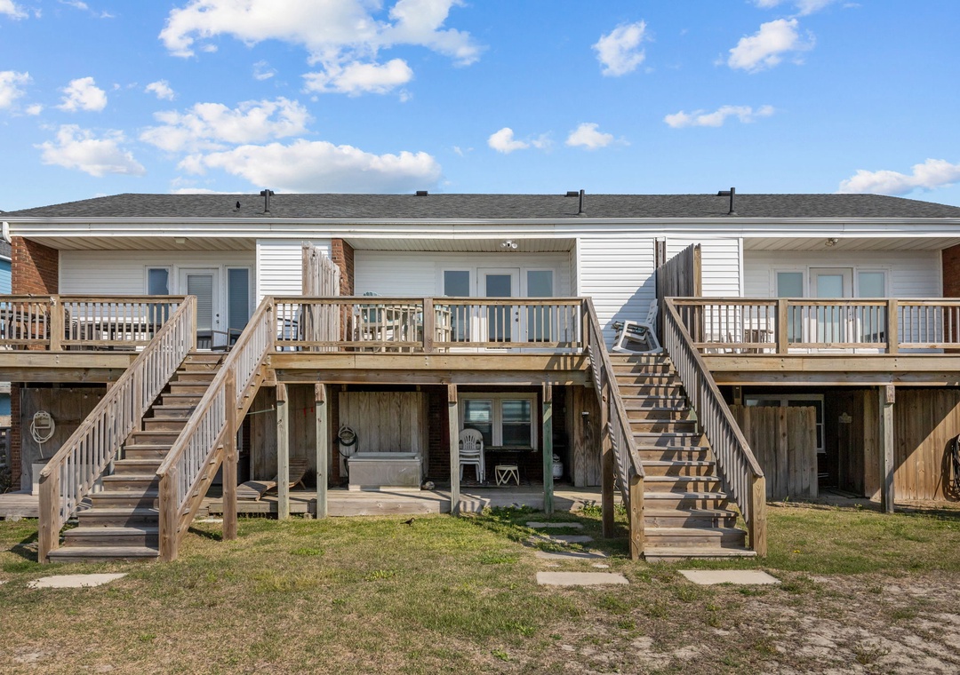 A beachside duplex with dual elevated decks and ground-level patios, surrounded by coastal grasses under bright blue skies.