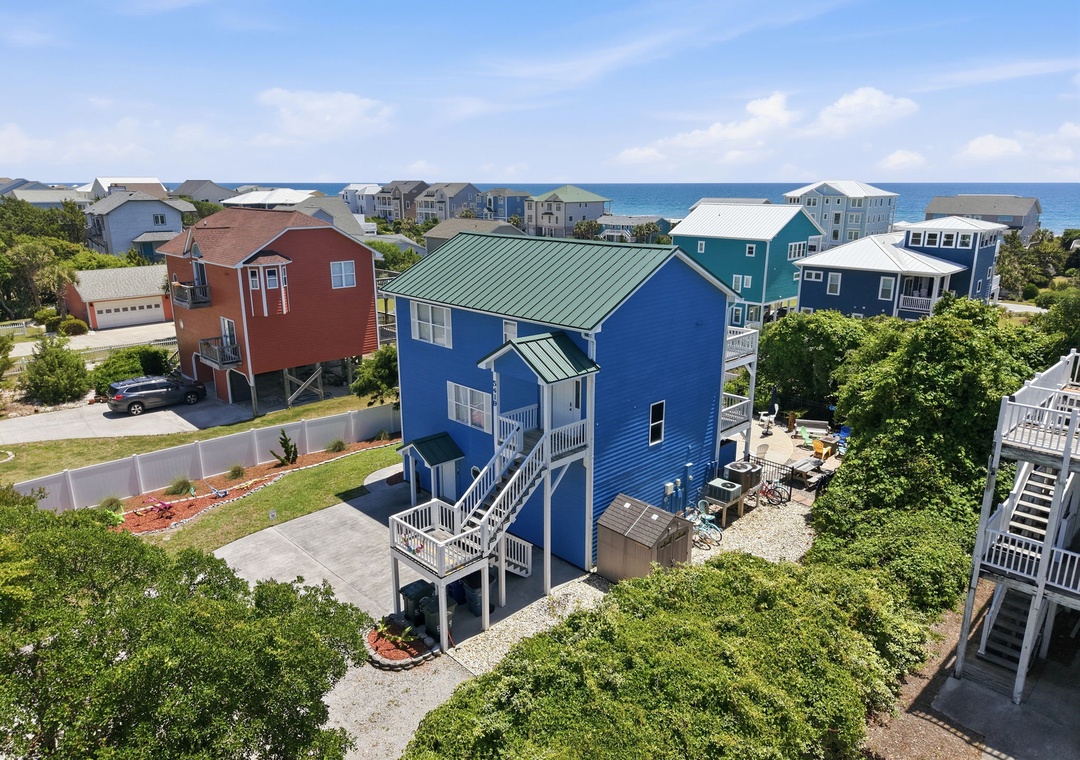 Aerial view of a bright blue coastal rental home featuring elevated construction, covered parking, outdoor patio space, and a quiet beachside neighborhood setting with ocean views in the distance.