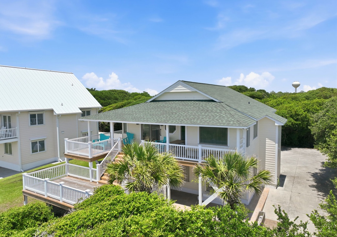 Elevated beach cottage with spacious wraparound decks and coastal landscaping, situated among lush greenery near the shore.