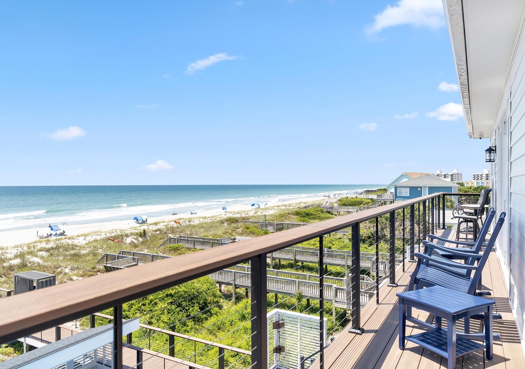 An expansive balcony overlooking the Atlantic coastline, complete with Adirondack chairs for leisurely mornings and sunset views. Sandy shores and coastal greenery stretch below.
