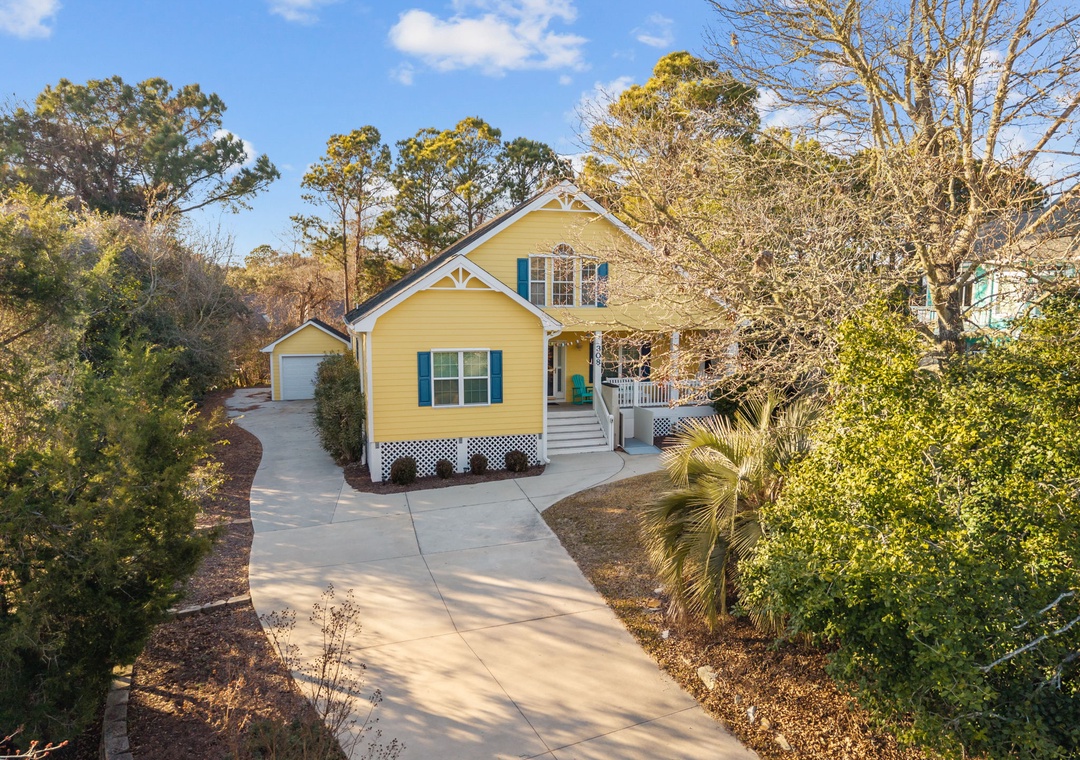 Charming yellow cottage nestled among towering trees and mature landscaping, featuring a welcoming front porch and peaceful residential setting.