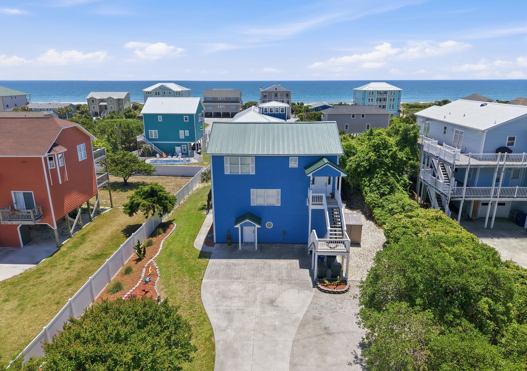 Aerial view of a blue coastal property situated in a quiet neighborhood, just steps from the ocean, surrounded by lush greenery and nearby vacation homes.