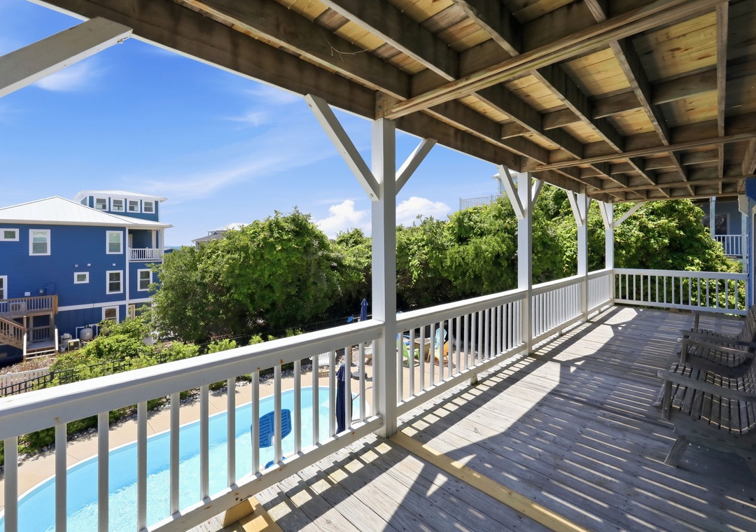 A covered deck featuring poolside views and glimpses of neighboring beach houses, with wicker seating to enjoy this sunny coastal setting.