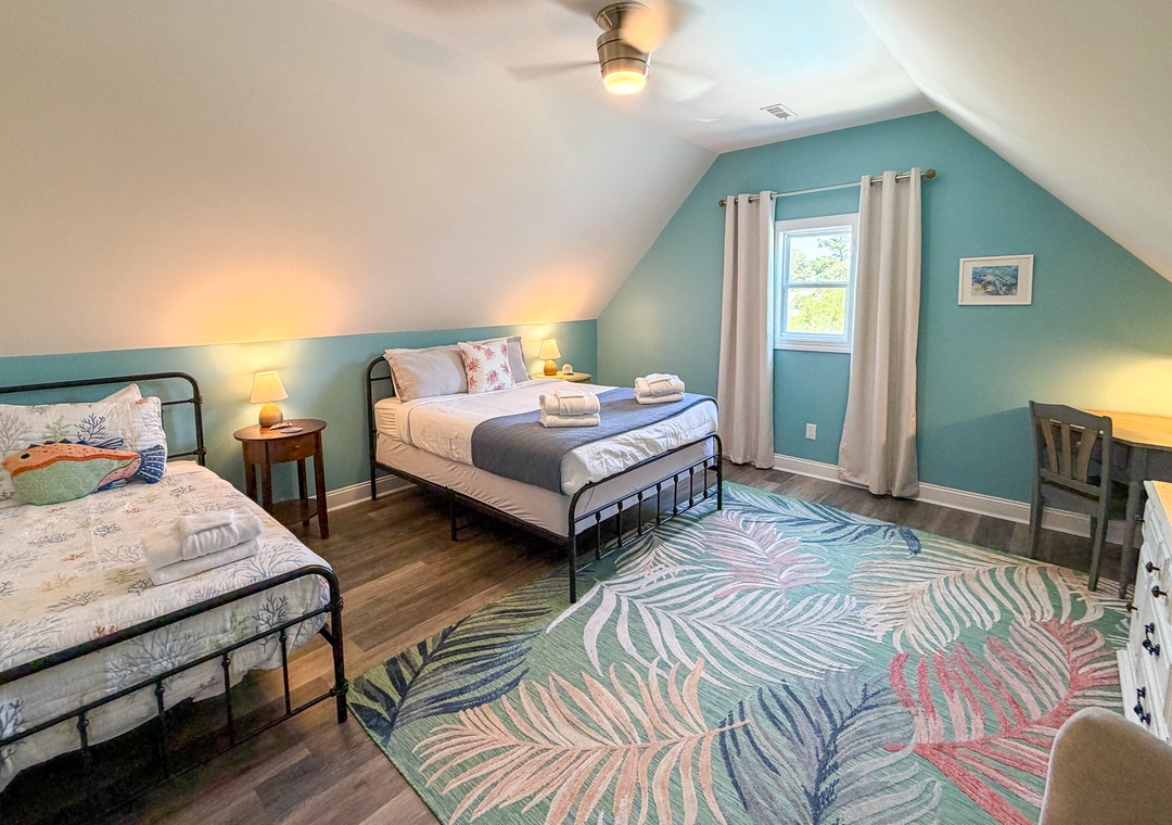 Your cozy retreat awaits with two comfortable beds, soothing teal walls, a ceiling fan for comfort, and warm natural light flowing through the window. A peaceful space to recharge and relax.
